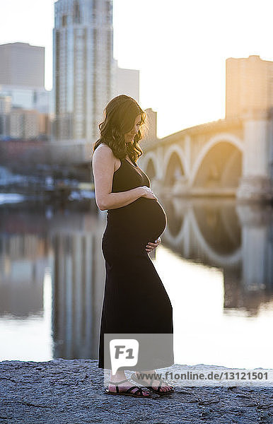 Pregnant woman standing by river against clear sky in city during sunset
