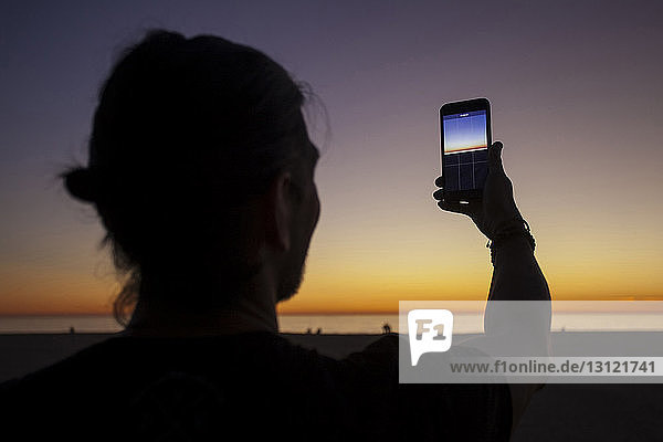 Nahaufnahme eines Mannes  der mit einem Smartphone vor dramatischem Himmel am Strand fotografiert