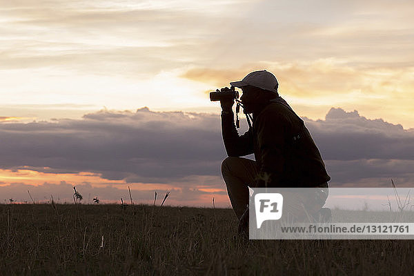 Seitenansicht eines Mannes  der ein Fernglas benutzt  während er bei Sonnenuntergang auf einem Feld im Serengeti-Nationalpark kniet