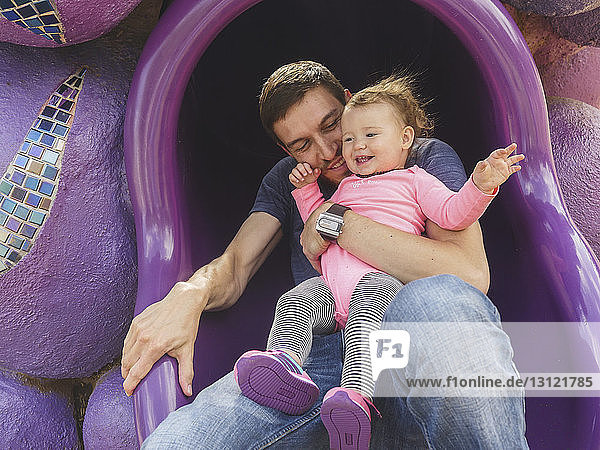 Vater mit süßer Tochter spielt auf dem Spielplatz auf einer Röhrenrutsche