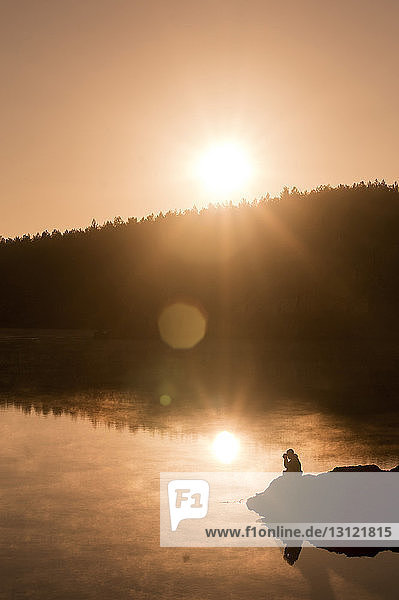 Silhouettenmann sitzt bei Sonnenuntergang am See gegen den Himmel