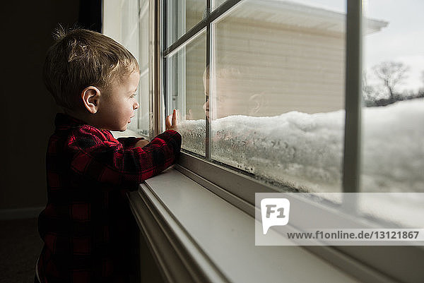 Seitenansicht eines Jungen  der durch ein Fenster schaut  während er im Winter zu Hause steht