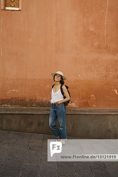 Porträt einer selbstbewussten Touristin in voller Länge mit Rucksack und Händen in den Taschen  die in der Stadt an der Wand steht