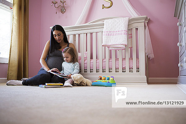 Schwangere Frau sitzt mit Tochter gegen Kinderbett zu Hause