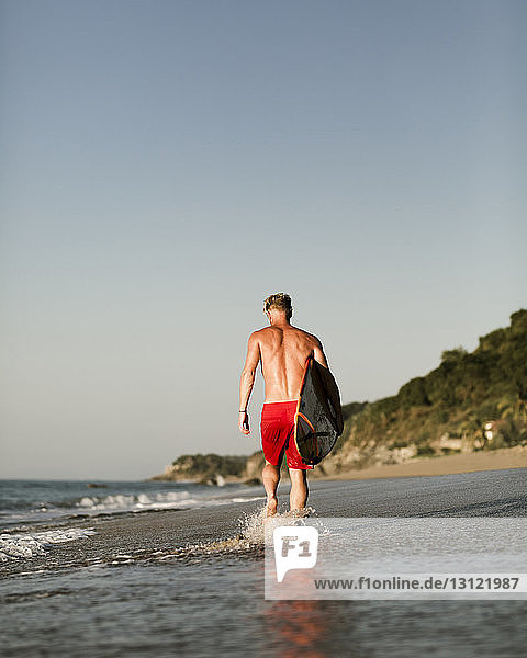 Rückansicht eines Mannes ohne Hemd mit Surfbrett  der am Strand vor klarem Himmel spazieren geht