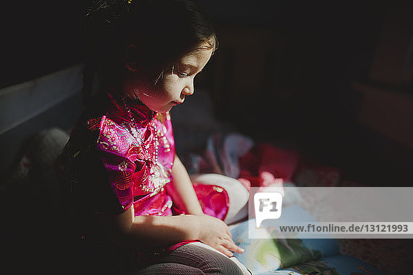 Girl looking at picture book while sitting on bed at home in darkroom