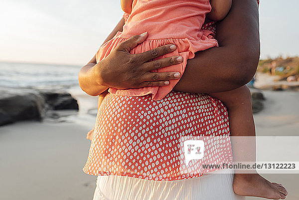 Mittelsektion einer schwangeren Mutter  die eine Tochter trägt  während sie bei Sonnenuntergang am Strand steht