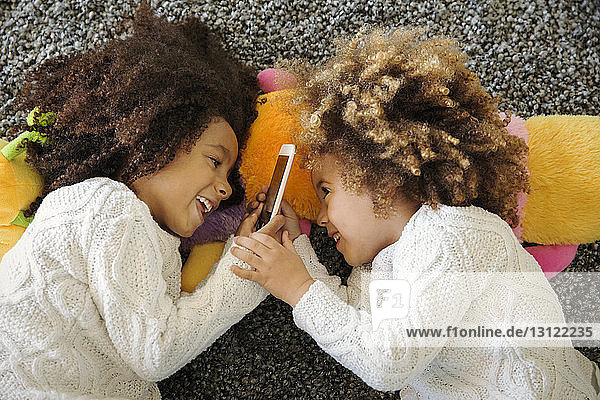 Hochwinkelansicht von Schwestern  die auf dem Teppich liegend mit einem Smartphone spielen