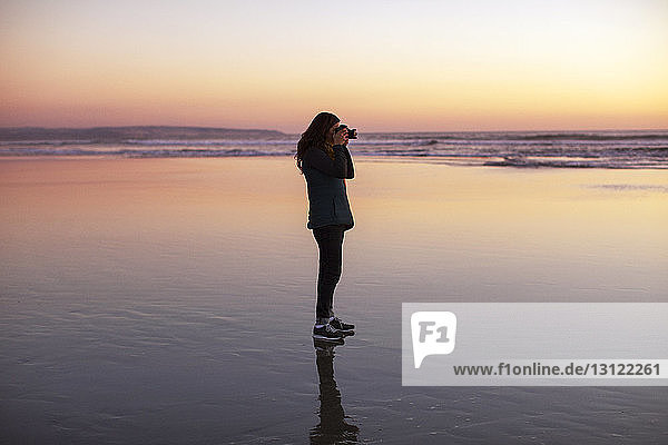 Seitenansicht einer Frau  die bei Sonnenuntergang am Strand steht und fotografiert