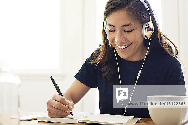 Cheerful woman listening music while writing in book at home
