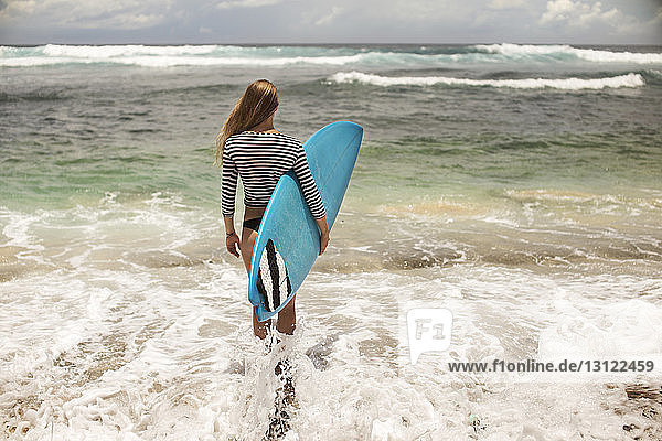 Rückansicht einer Frau  die ein Surfbrett trägt  während sie im Meer steht