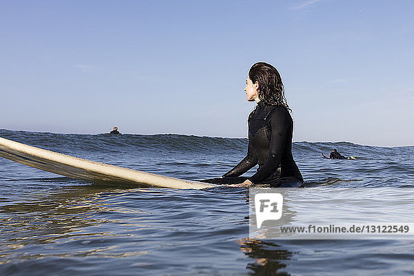 Nachdenkliche Surferin sitzt auf einem Surfbrett im Meer vor klarem Himmel