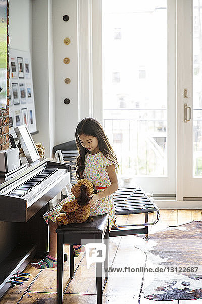 Mädchen sitzt mit Teddybär am Klavier zu Hause