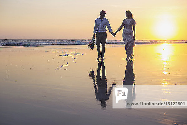 Frisch verheiratetes Paar hält sich bei Sonnenuntergang beim Strandspaziergang am klaren Himmel an den Händen