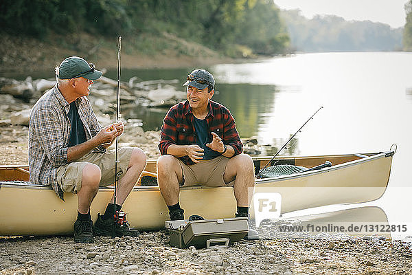 Male friends with fishing rods talking while sitting on boat at lakeshore