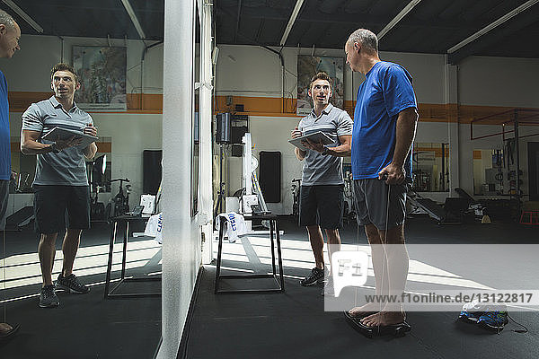 Trainer mit Tablet-Computer im Gespräch mit einem Mann  der im Fitnessstudio Dehnübungen macht