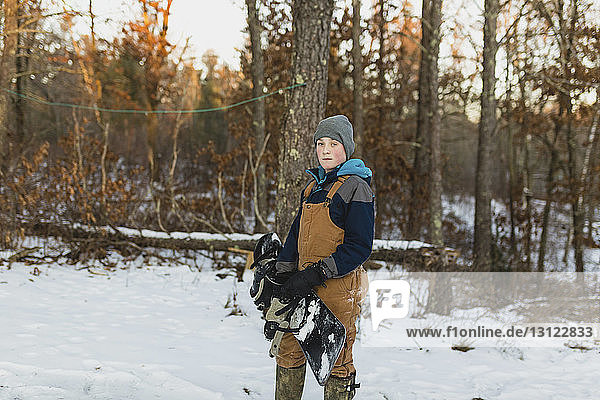 Portrait of boy with snowboard standing on snow covered field in forest