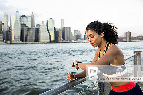 Female athlete checking smart watch while standing by river against sky in city