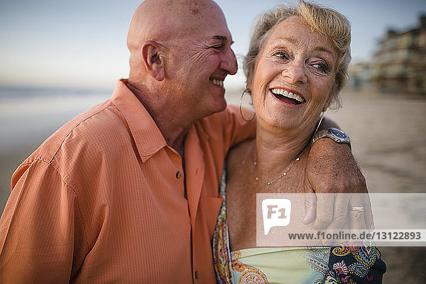 Happy senior couple standing at beach against sky during sunset