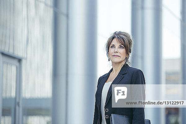 Confident businesswoman looking away while standing against office building
