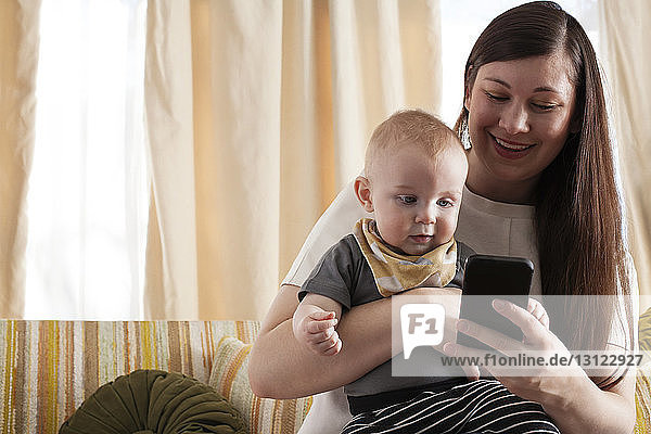 Happy mother showing mobile phone to baby while sitting on sofa at home