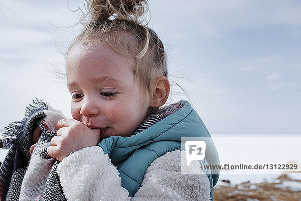 Close-up of crying girl sucking thumbs while standing against sky during winter