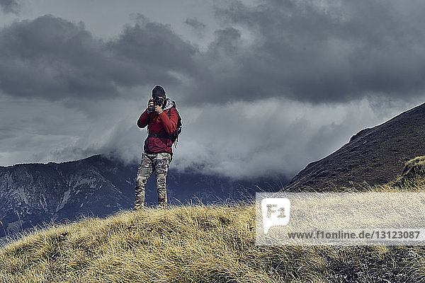 Male hiker photographing while standing on mountain against cloudy sky