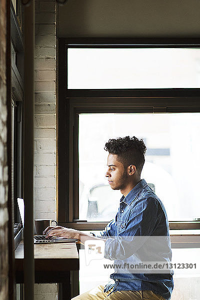 Seitenansicht eines Mannes  der seinen Laptop bei einer Kaffeetasse benutzt  während er an den Fenstern eines Cafés sitzt