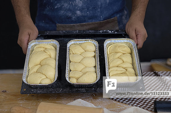 Mittelsektion eines Mannes  der in der Bäckerei stehend Brote in Behältern auf einem Backblech hält