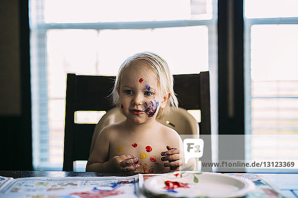 Boy with paint on face looking away while sitting at table