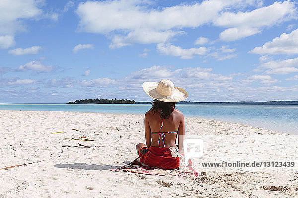 Rückansicht einer Frau mit Sonnenhut  die am Strand gegen den Himmel sitzt