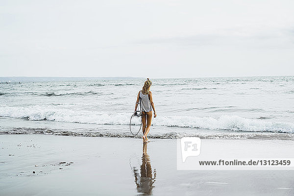 Rückansicht einer Frau  die ein Surfbrett trägt  während sie in Richtung Meer gegen den Himmel läuft