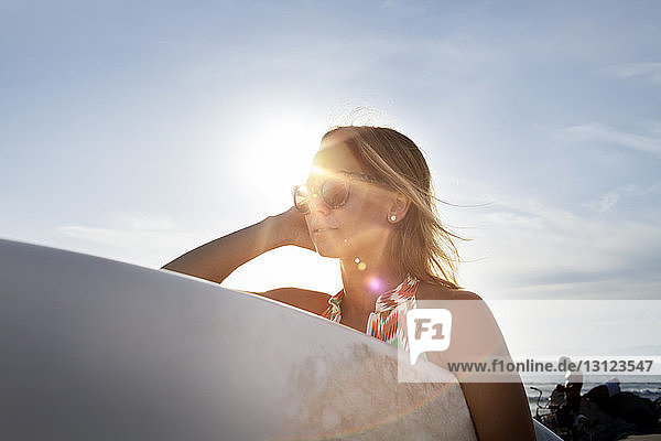 Nachdenkliche Frau trägt Surfbrett am Strand gegen den Himmel