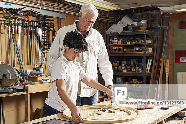 Carpenter looking at boy measuring wooden ring in workshop