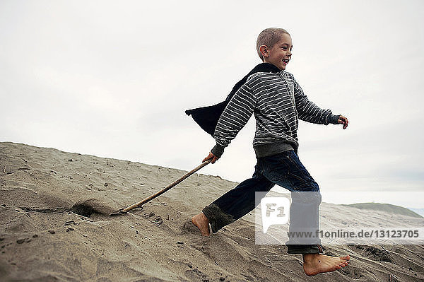 Verspielter Junge mit Stock  der auf Sand am Strand gegen den Himmel läuft