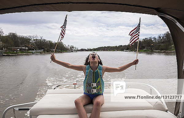 Glückliches Mädchen schreit  während es amerikanische Flaggen im Boot auf dem See gegen den bewölkten Himmel hält