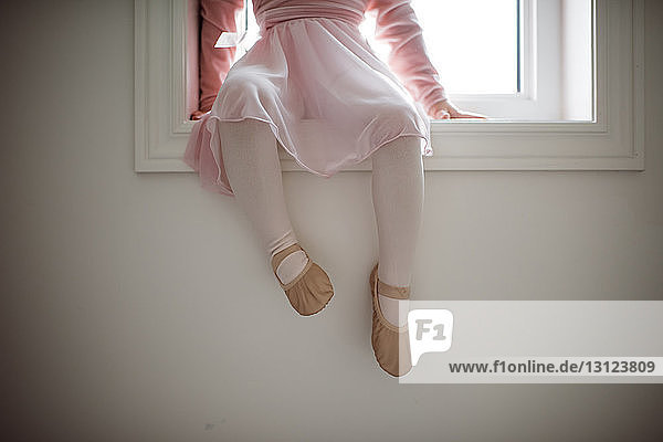 Low section of girl in ballet costume sitting on window sill at home
