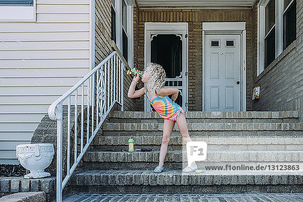 Verspieltes Mädchen bläst Spielzeugpfeife  während es auf Stufen gegen das Haus steht