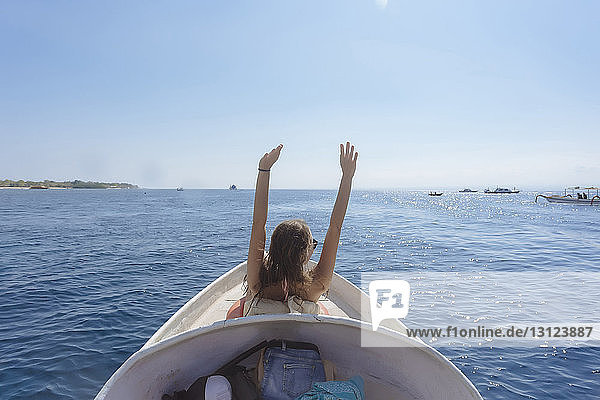 Rückansicht einer Frau mit erhobenen Armen  die im Boot auf dem Meer vor klarem Himmel steht