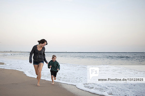 Mother and son running on shore at beach against sky during sunset