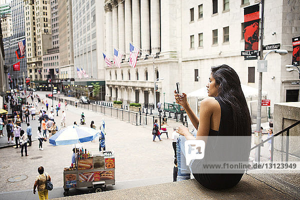 Frau fotografiert über Mobiltelefon  während sie auf Stufen in der Stadt sitzt