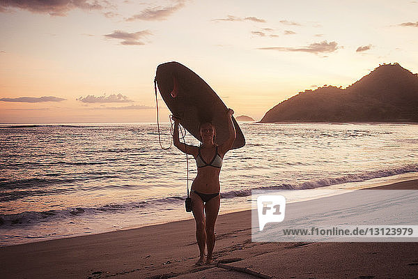 Porträt einer Surferin  die bei Sonnenuntergang am Strand ein Surfbrett auf dem Kopf trägt