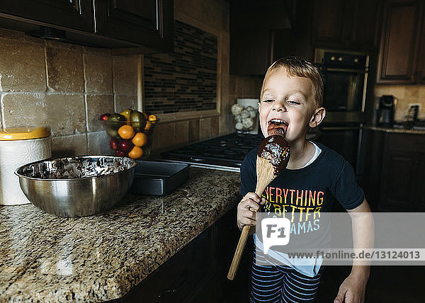 Glücklicher Junge leckt Löffel  während er in der Küche steht