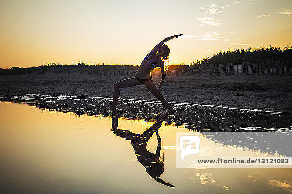 Frau im Bikini  die bei Sonnenuntergang am Strand Yoga praktiziert