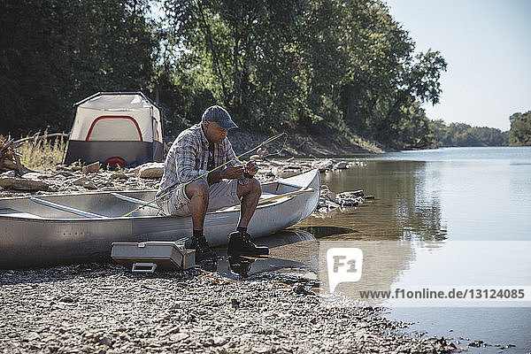Man adjusting fishing tackle while sitting on boat at lakeshore