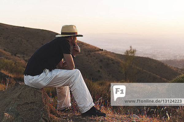 Nachdenklicher Mann schaut weg  während er bei Sonnenuntergang auf einem Felsen gegen klaren Himmel und Berge sitzt