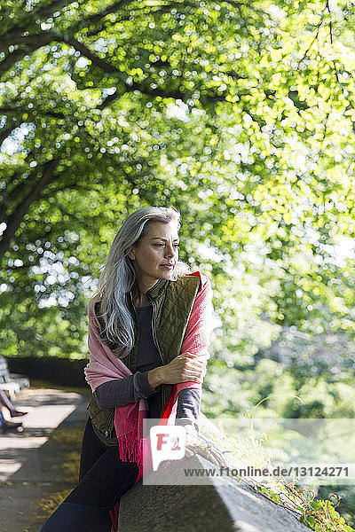 Thoughtful woman looking away while leaning on retaining wall against branches