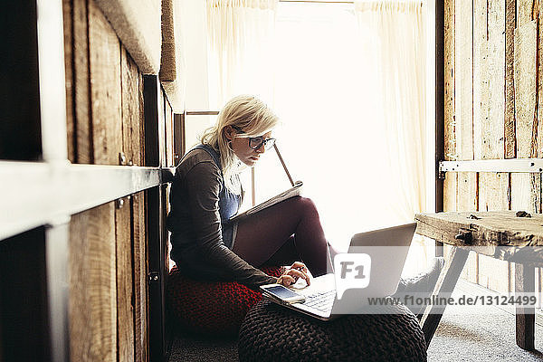 Junge Geschäftsfrau benutzt Laptop  während sie im Büro auf einem Ottoman sitzt