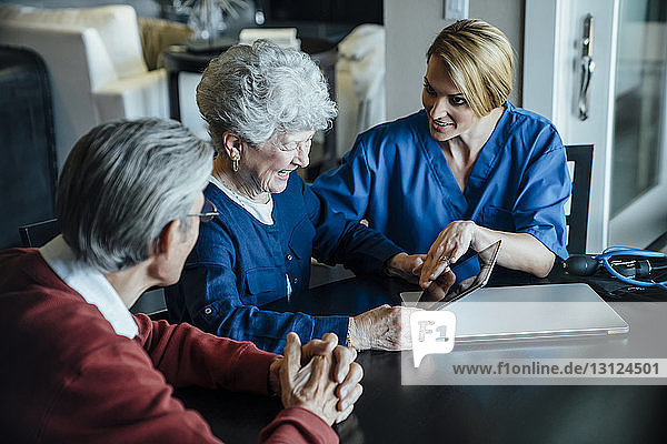Heimpflegekraft diskutiert am Esstisch über Tablet-Computer mit älterem Ehepaar