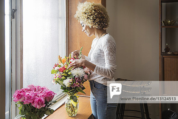 Lächelnde junge Frau arrangiert Blumen in Container auf der Fensterbank zu Hause
